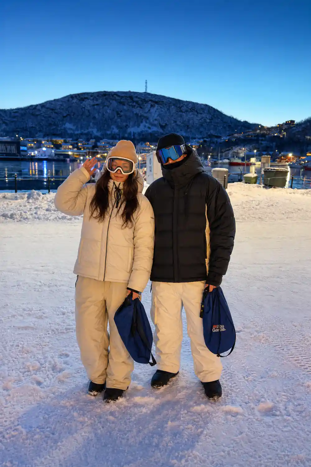 Two travellers in flotation suits and ski goggles on shore, harbour behind